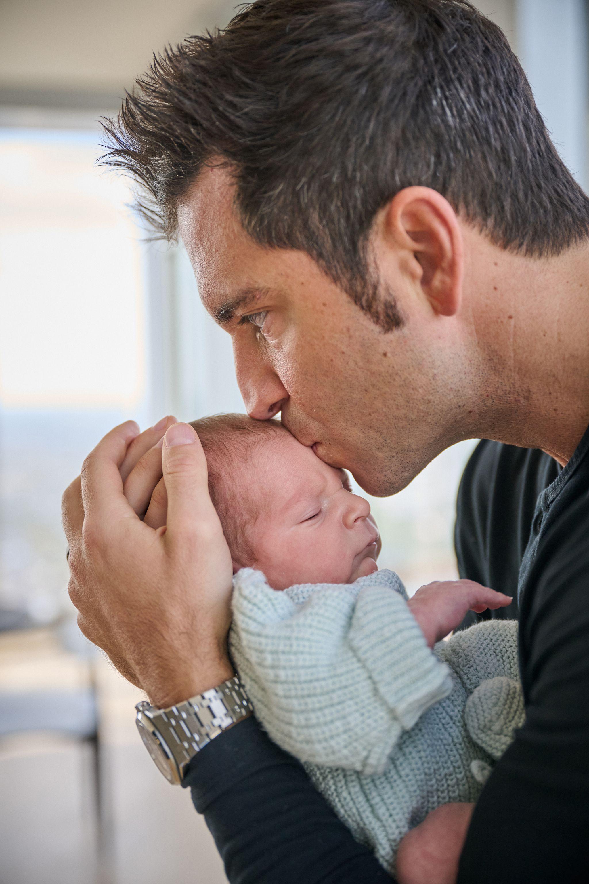 Father cradling his newborn and kissing the baby's head beside a high window — Newborn photography, Austin Newborn