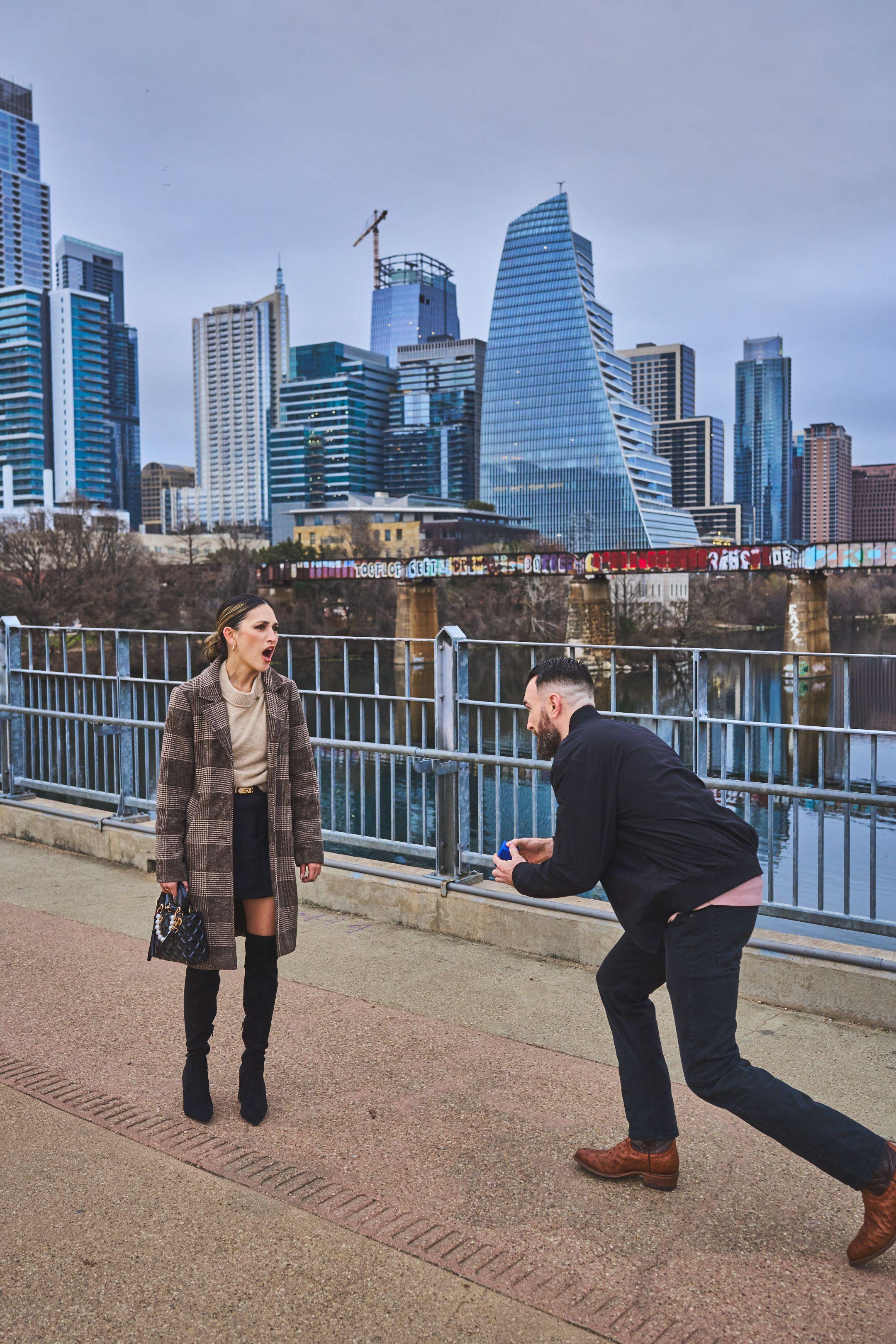 Woman covering her face as her partner begins to kneel on the bridge, skyline — Engagement & Proposals photography, Austin A woman covering her face as her partner begins to kneel on the bridge, skyline behind.