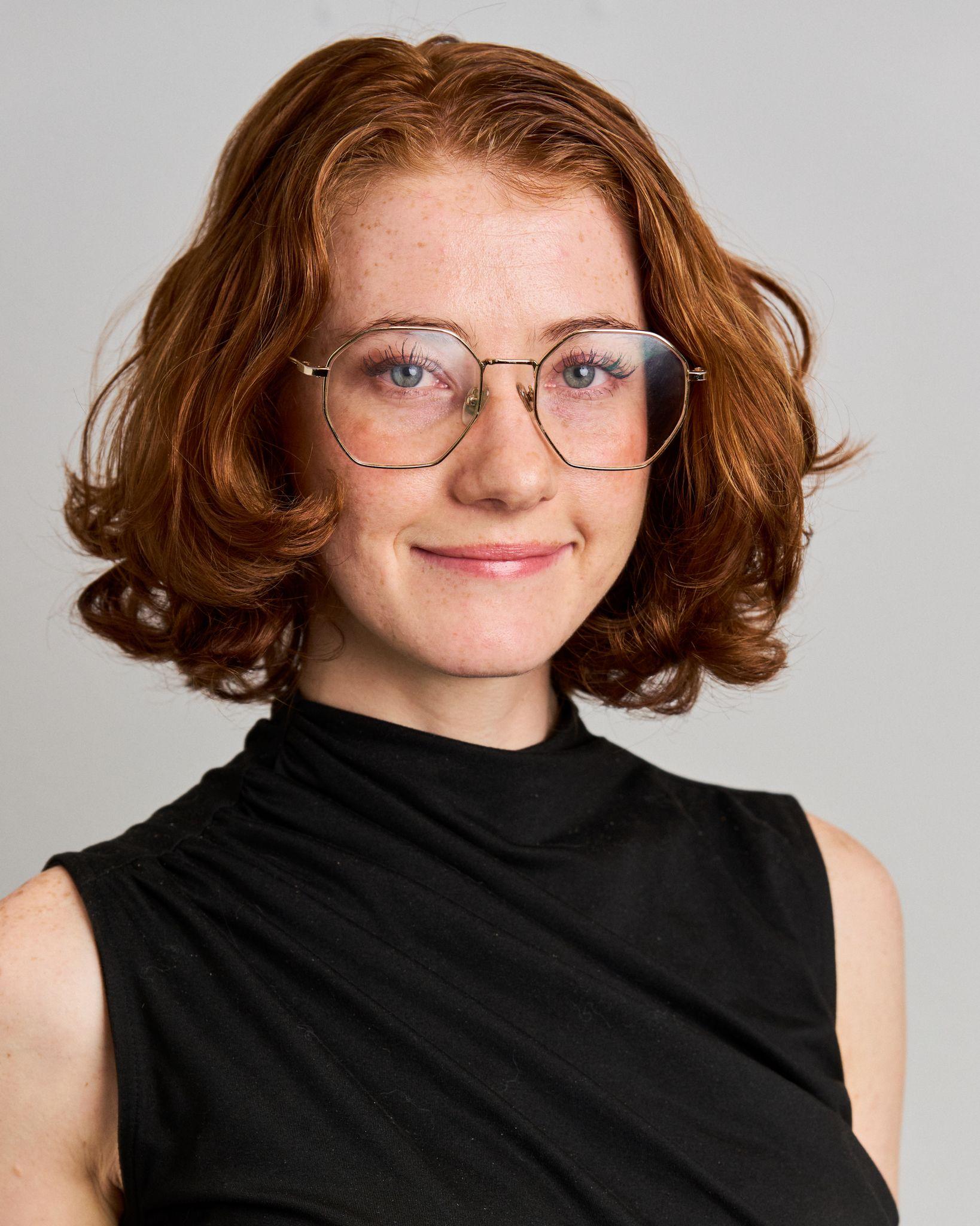 A red-haired woman in wire-framed glasses and a black sleeveless turtleneck against a gray studio background