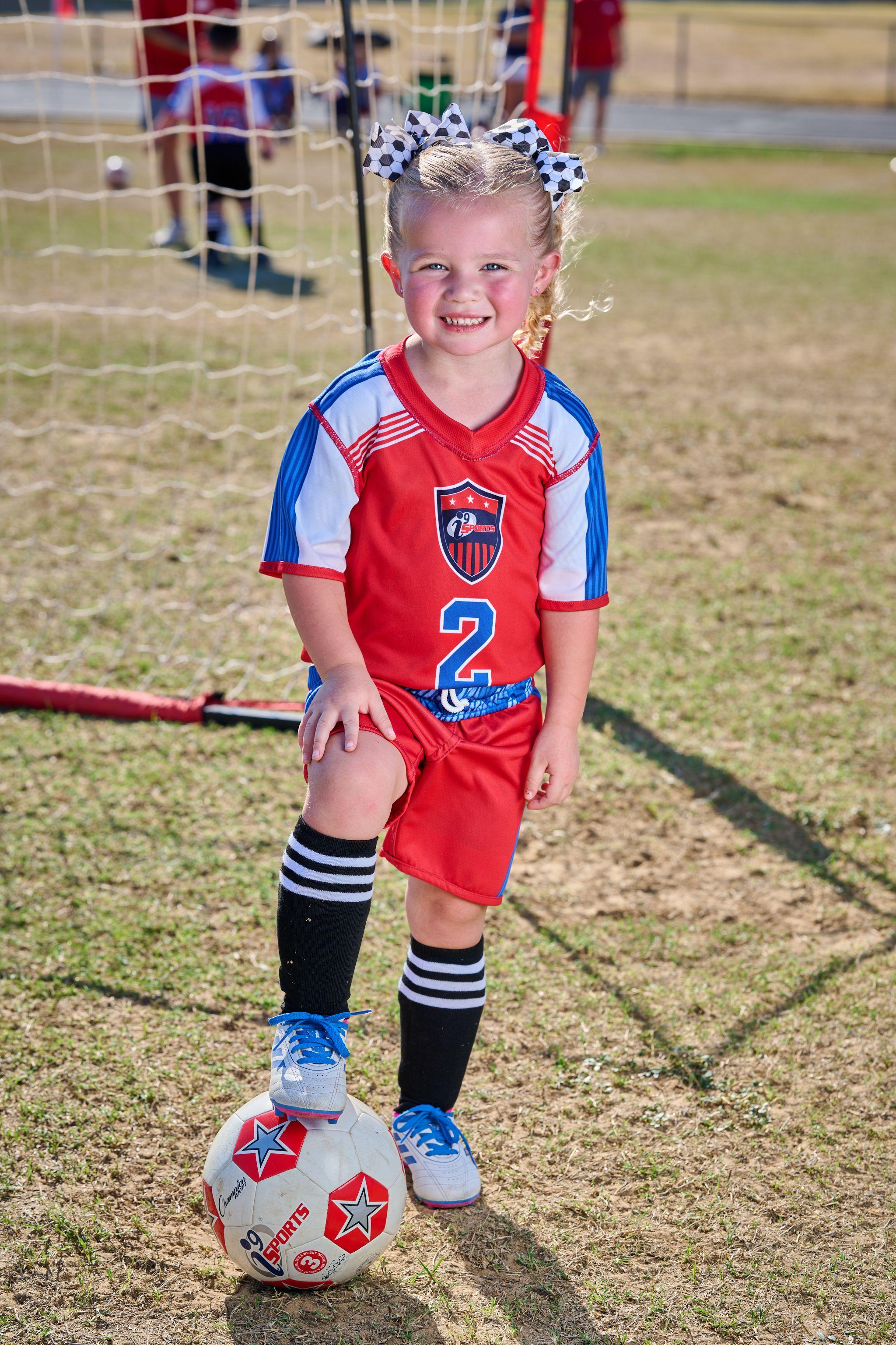 Small child in a red kit standing with one foot on a football beside a goal — School event photography, Austin Small child in a red kit standing with one foot on a football beside a goal