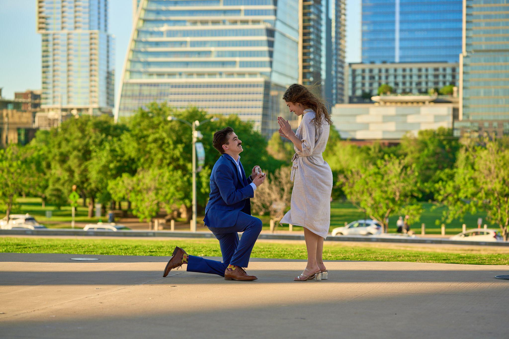 Man kneeling to propose on a park path as his partner covers her face, towers — Butler Metro Park photography, Austin A man kneeling to propose on a park path as his partner covers her face, towers behind.