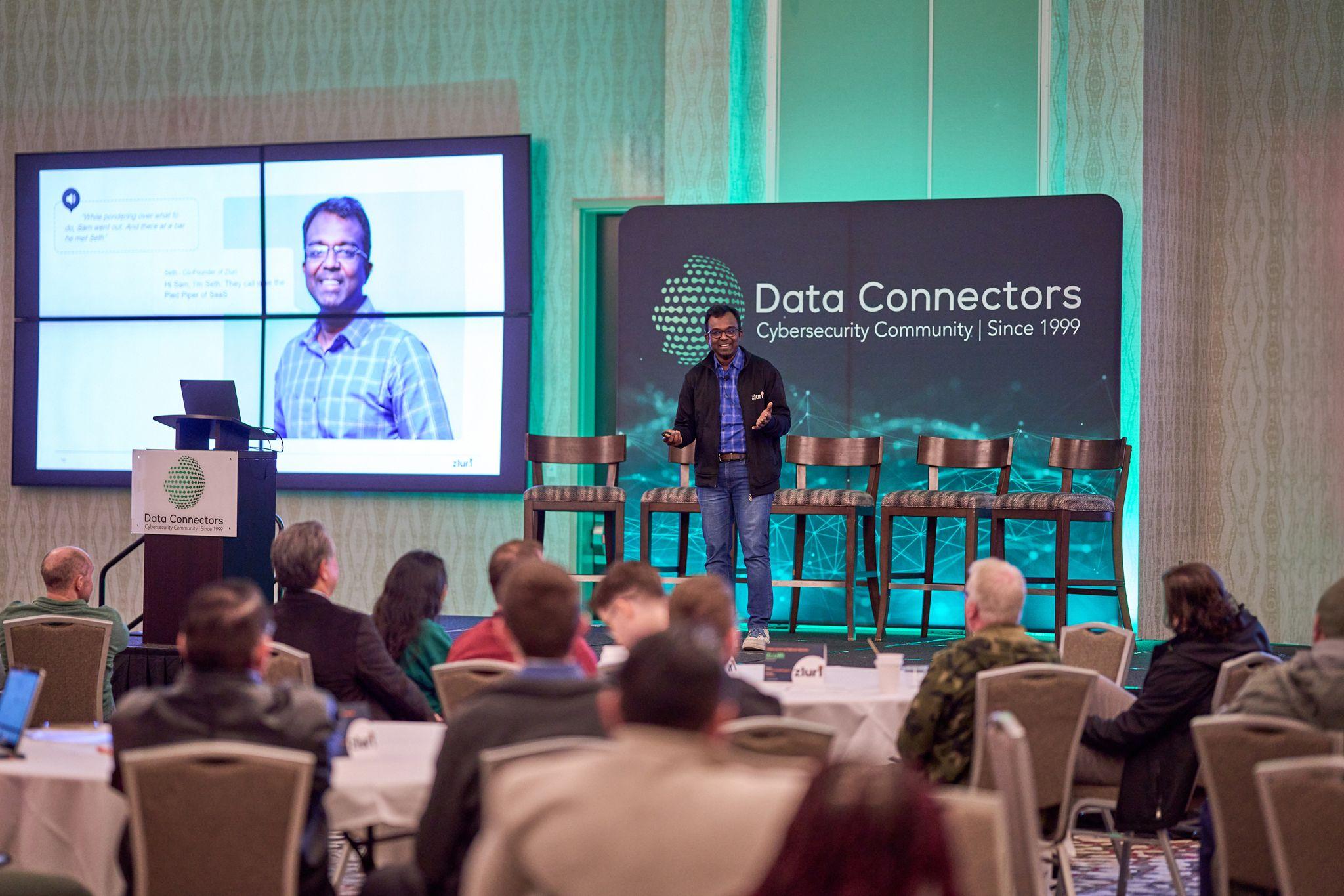 Speaker presenting on a Data Connectors cybersecurity conference stage — Conventions & Tradeshows photography, Austin A speaker presenting on a Data Connectors cybersecurity conference stage above seated round tables.