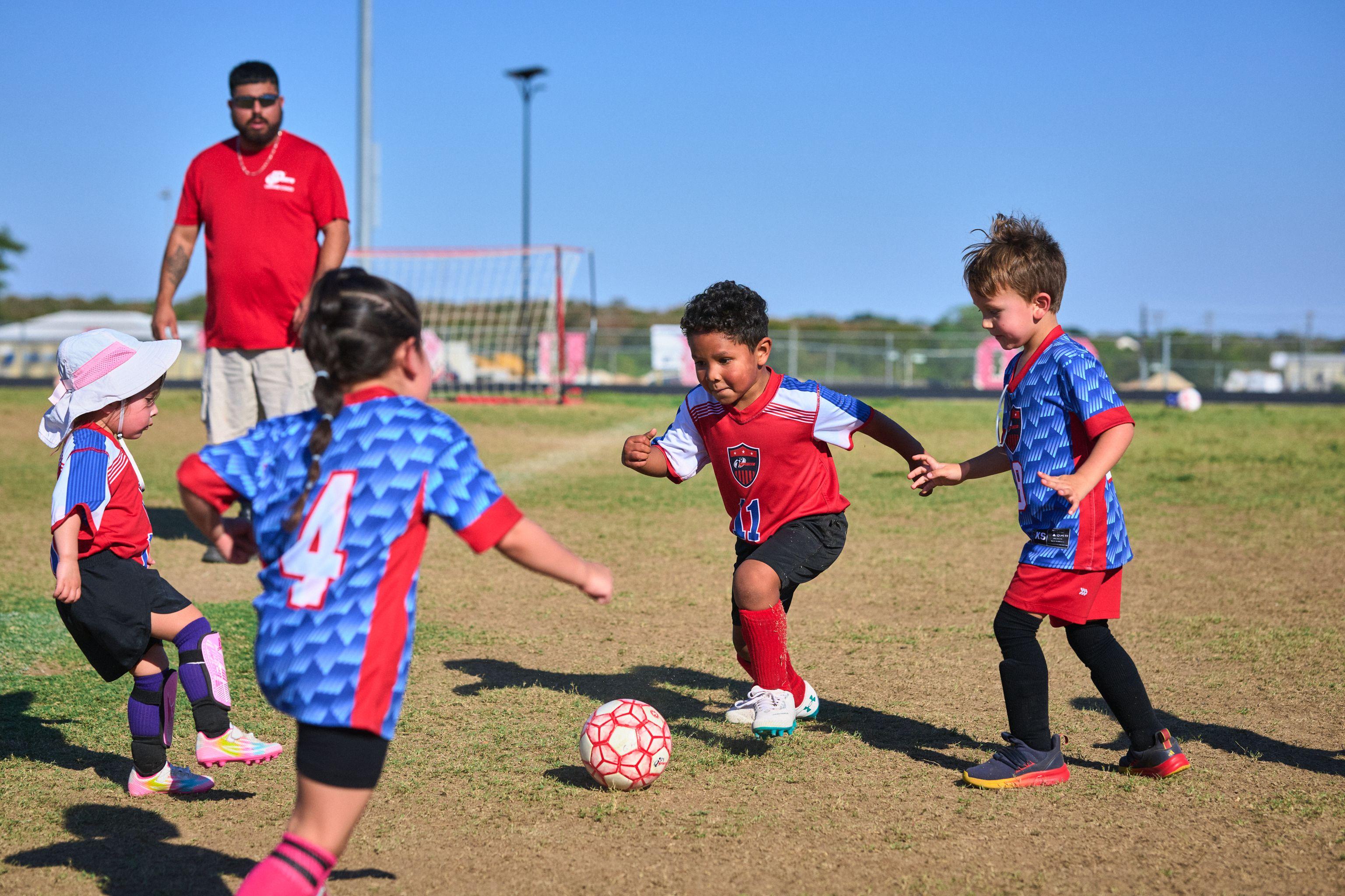 Group of young children in red and blue kits converging on the ball during a match — Sports photography, Austin Group of young children in red and blue kits converging on the ball during a match