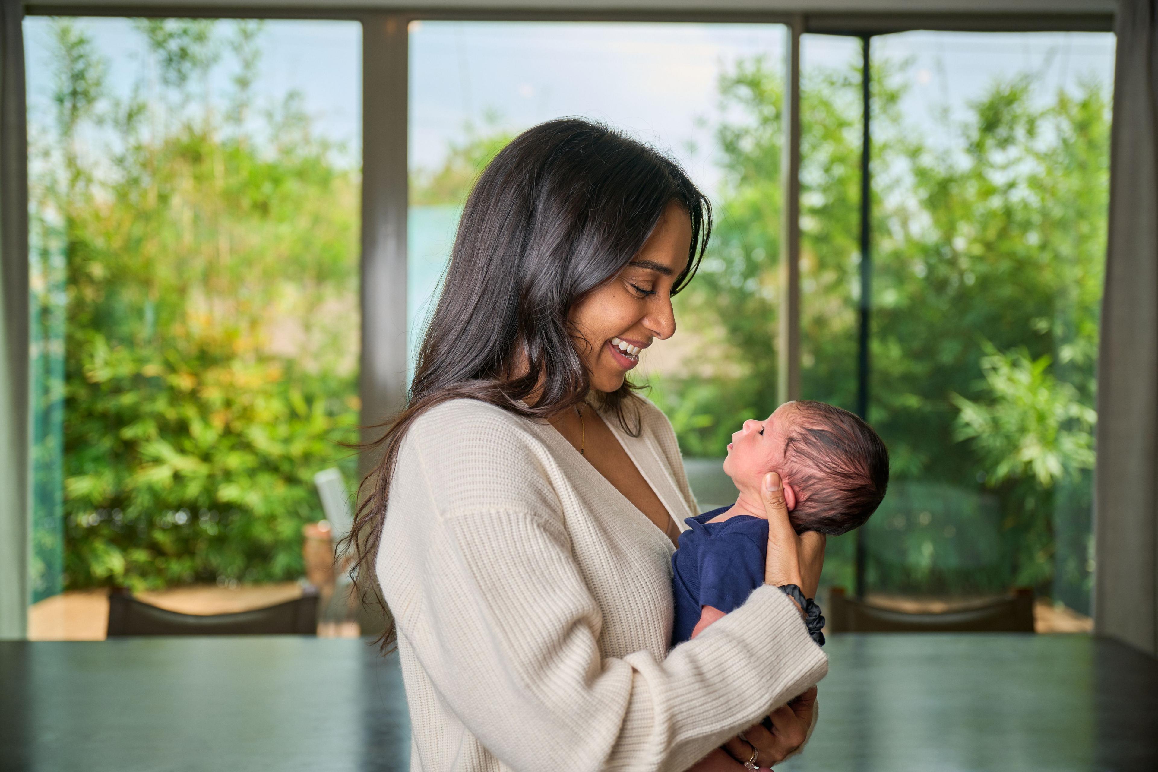Mother standing at a window holding her newborn, the garden outside softly out of focus — Mobile photo studio, Austin A mother standing at a window holding her newborn, the garden outside softly out of focus