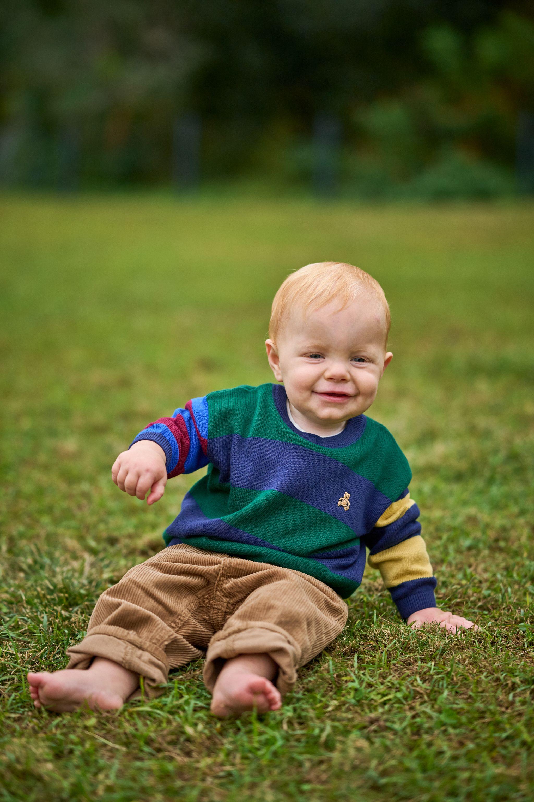Toddler in a striped jumper sitting on the grass, mid-laugh — Waterloo Park photography, Austin A toddler in a striped jumper sitting on the grass, mid-laugh.