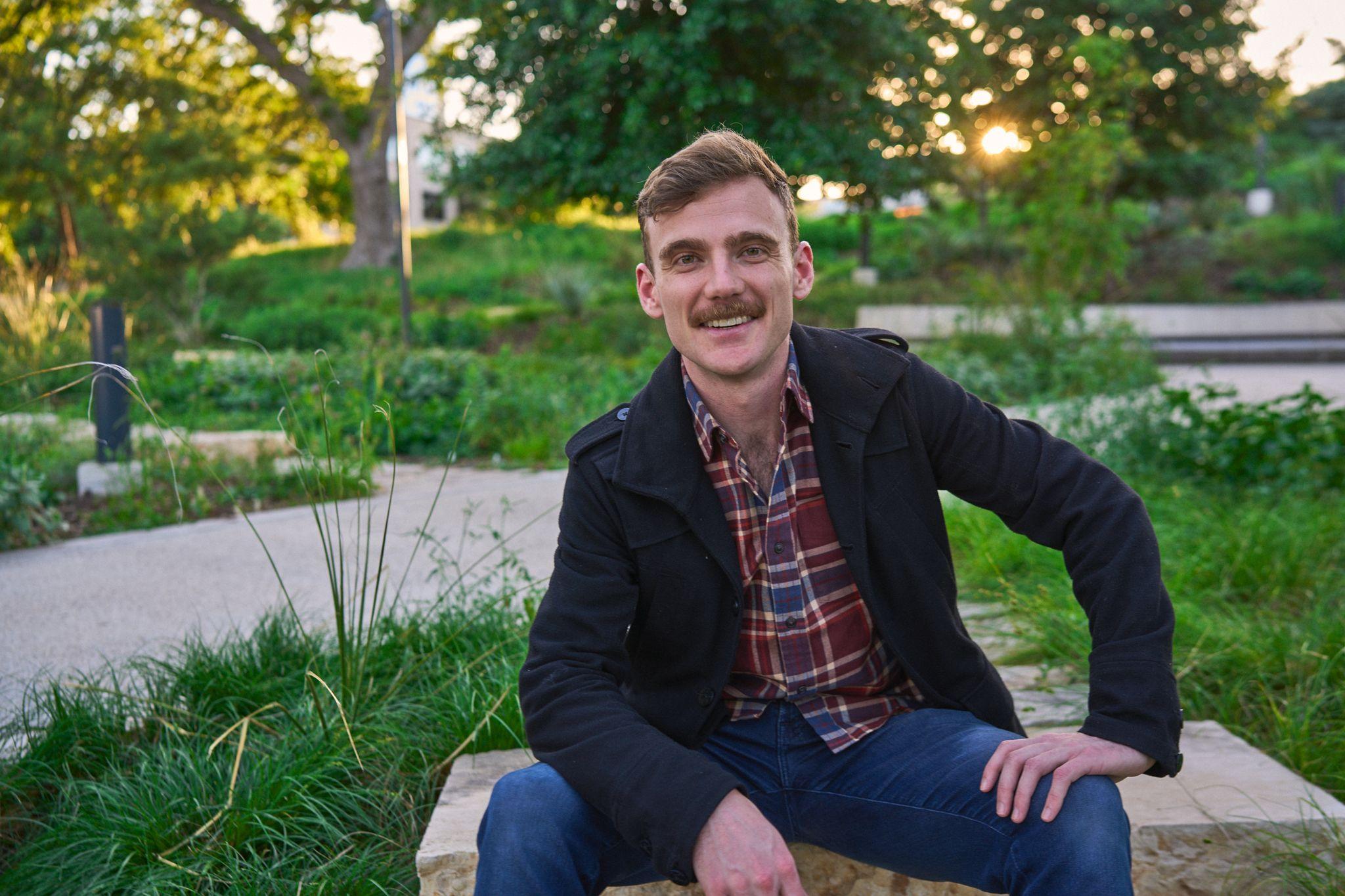 Man in a plaid shirt and dark jacket seated on a low wall at golden hour — Waterloo Park photography, Austin A man in a plaid shirt and dark jacket seated on a low wall at golden hour.