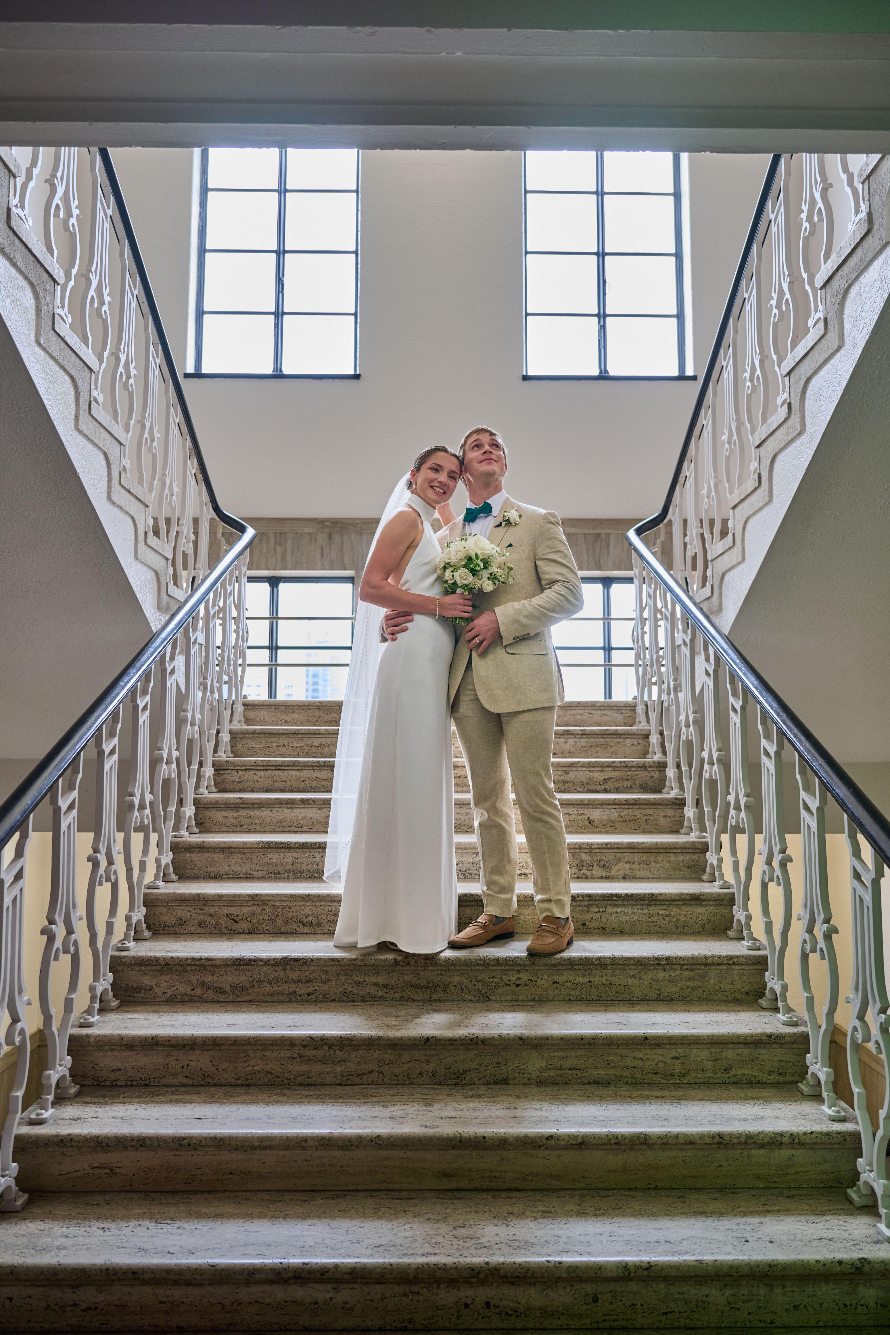 Couple standing at the top of a grand courthouse staircase framed by tall windows — Weddings photography, Austin A couple standing at the top of a grand courthouse staircase framed by tall windows.