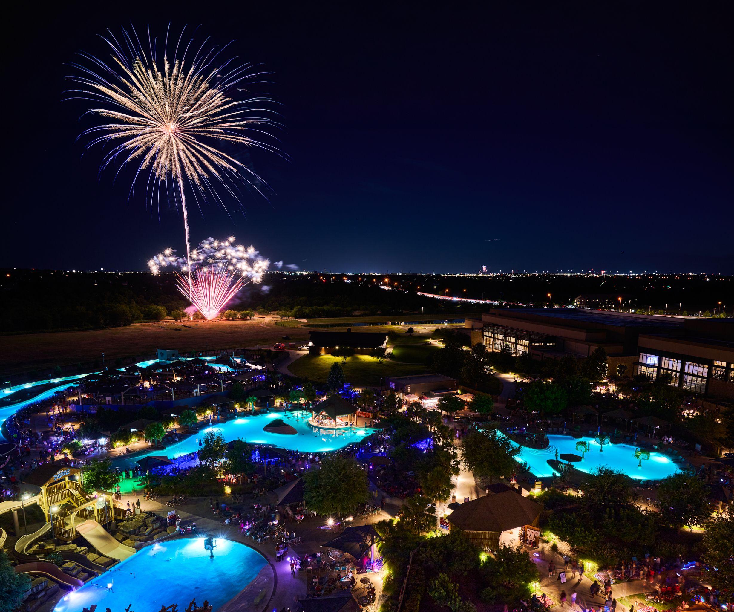 Fireworks bursting above a resort waterpark lit blue at night, seen from above — Hospitality & leisure photography, Austin Fireworks bursting above a resort waterpark lit blue at night, seen from above.