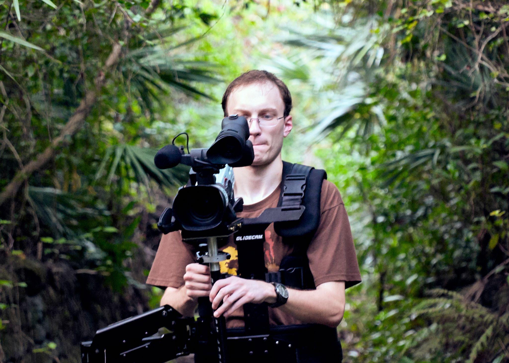 Camera operator with a shoulder-mounted cinema rig shooting from inside dense green foliage — Production behind-the-scenes, Austin Camera operator with a shoulder-mounted cinema rig shooting from inside dense green foliage