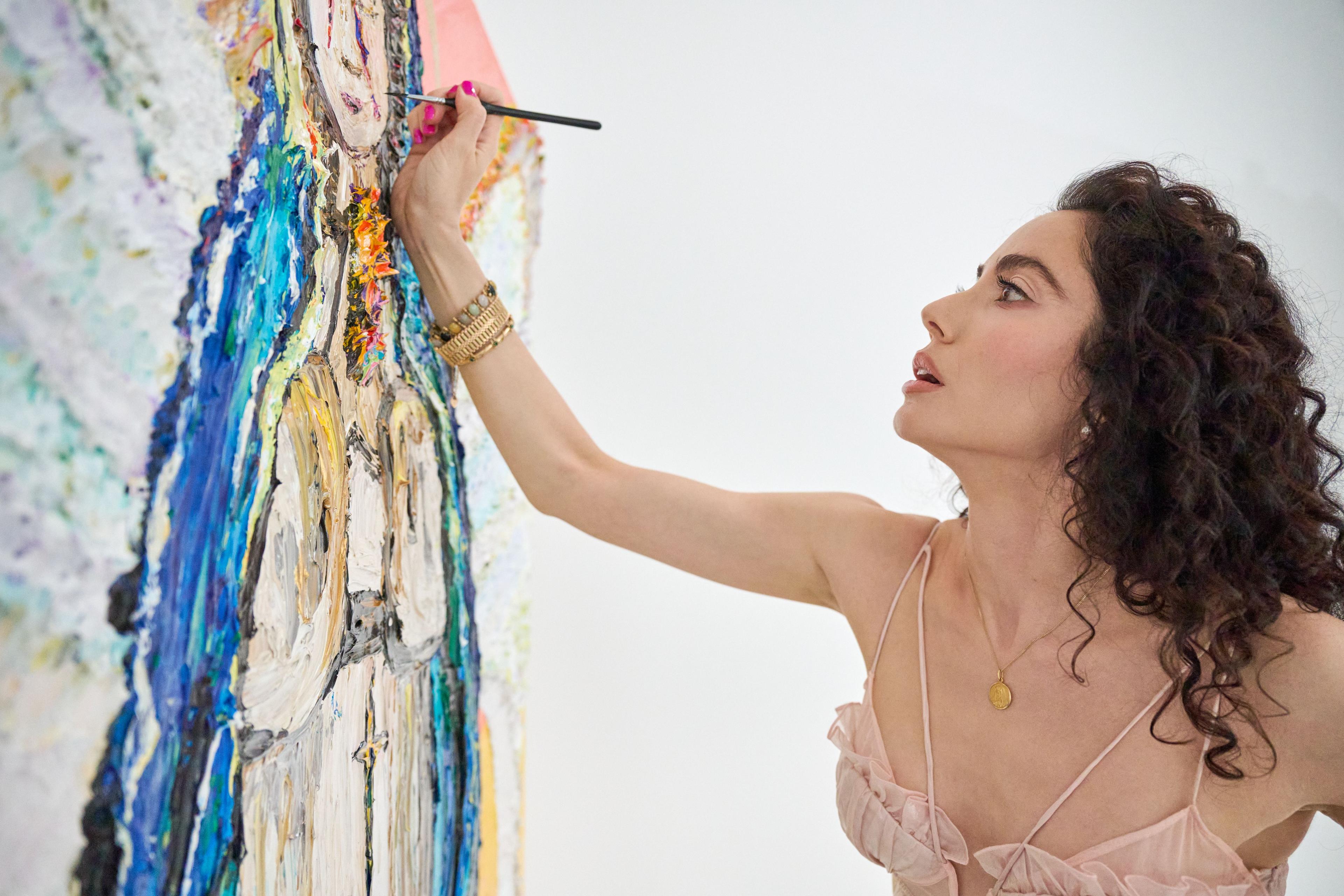Artist in a pale pink dress reaching up with a brush to paint a tall — Artwork & archival photography, Austin Artist in a pale pink dress reaching up with a brush to paint a tall, brightly colored figurative canvas