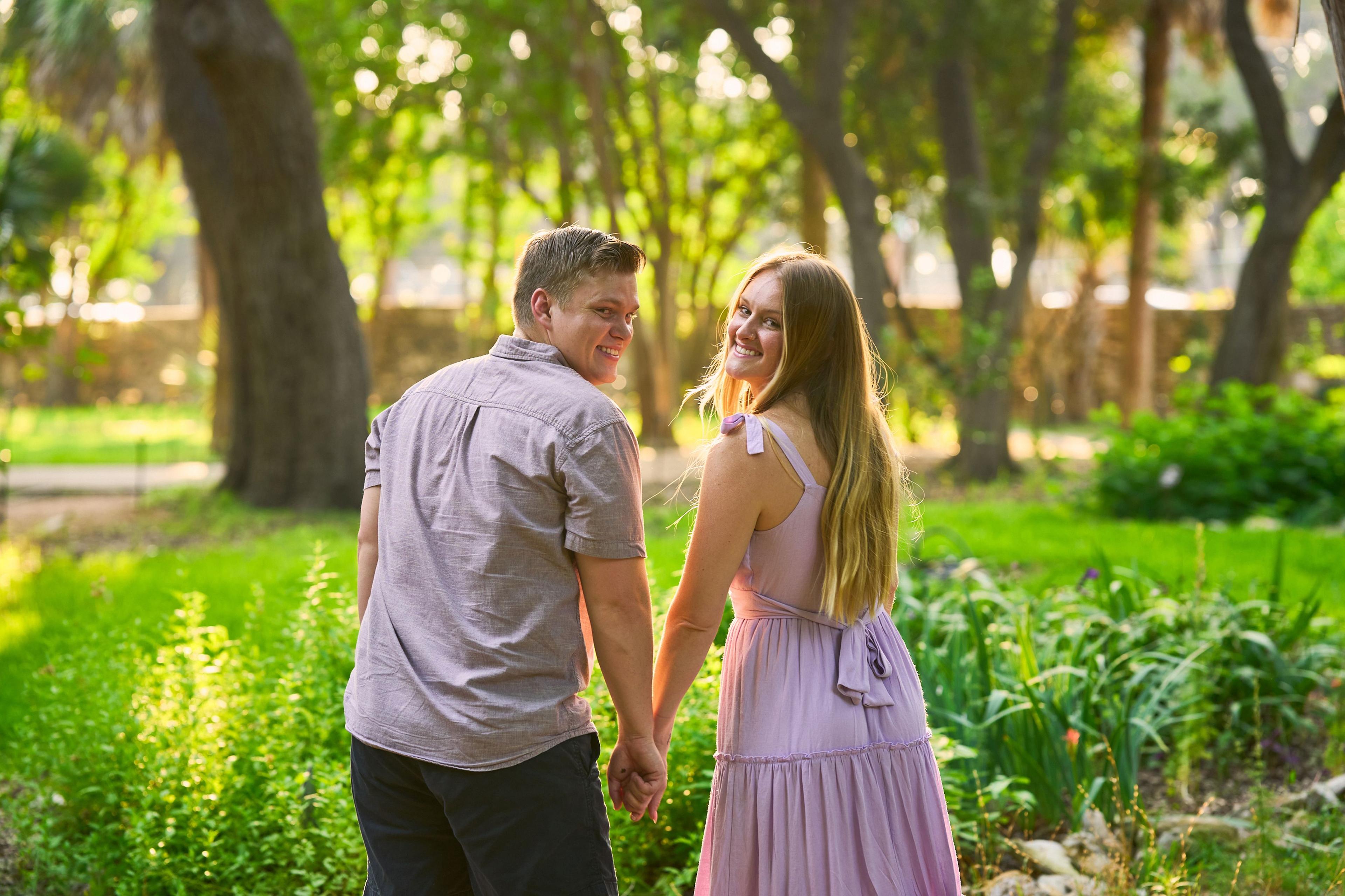 Couple walking away hand in hand down a green path at Mayfield Park, glancing back — Mayfield Park photography, Austin A couple walking away hand in hand down a green path at Mayfield Park, glancing back at the camera