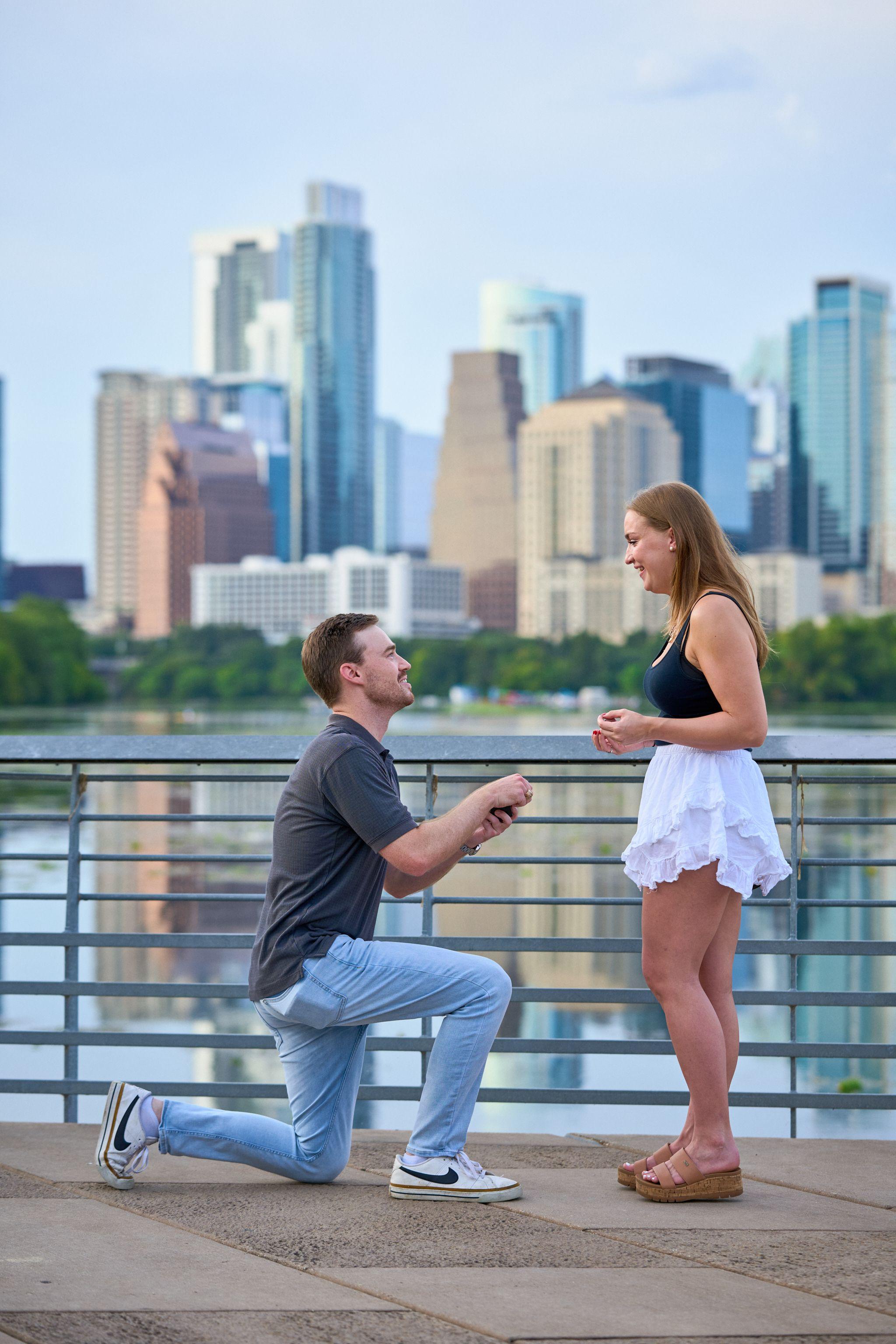 Man kneeling to propose on a boardwalk railing above Lady Bird Lake, downtown towers — Austin Boardwalk photography, Austin A man kneeling to propose on a boardwalk railing above Lady Bird Lake, downtown towers behind.