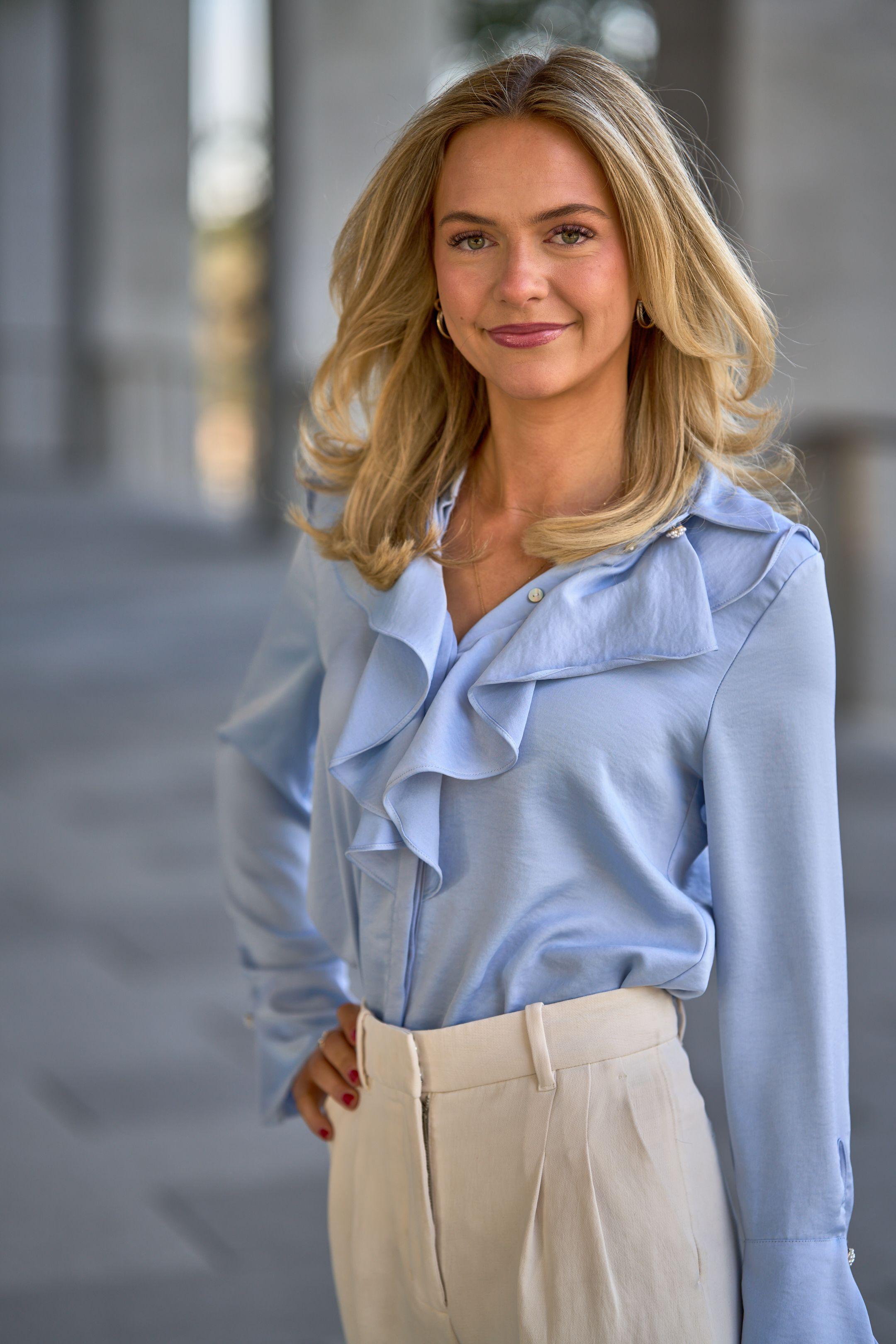 Woman in a pale blue ruffled blouse with one hand at her waist, photographed outdoors — Business headshots, Austin A woman in a pale blue ruffled blouse with one hand at her waist, photographed outdoors against a soft gray background