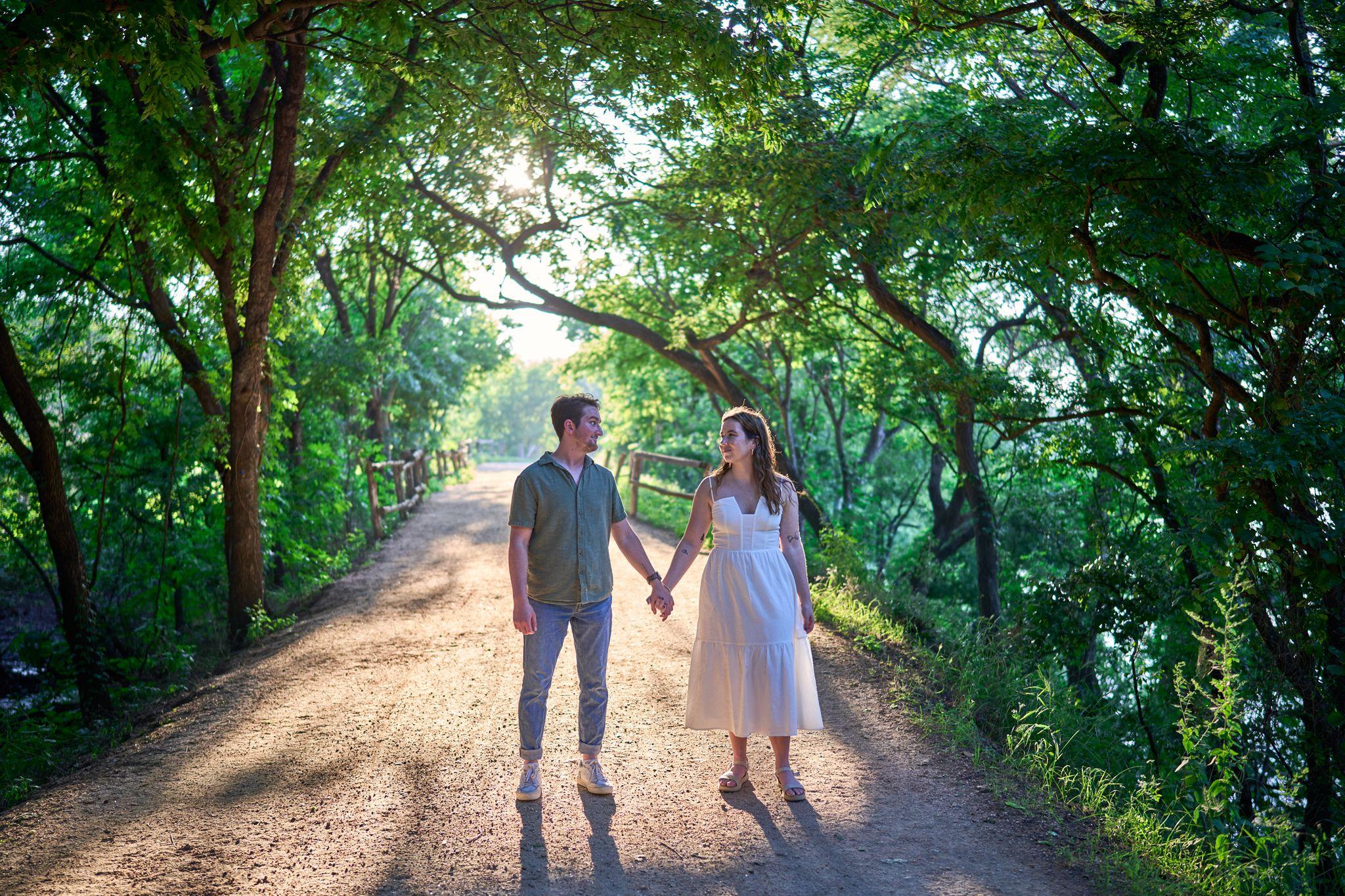 Couple holding hands on a shaded dirt path under a green canopy — Butler Metro Park photography, Austin A couple holding hands on a shaded dirt path under a green canopy.