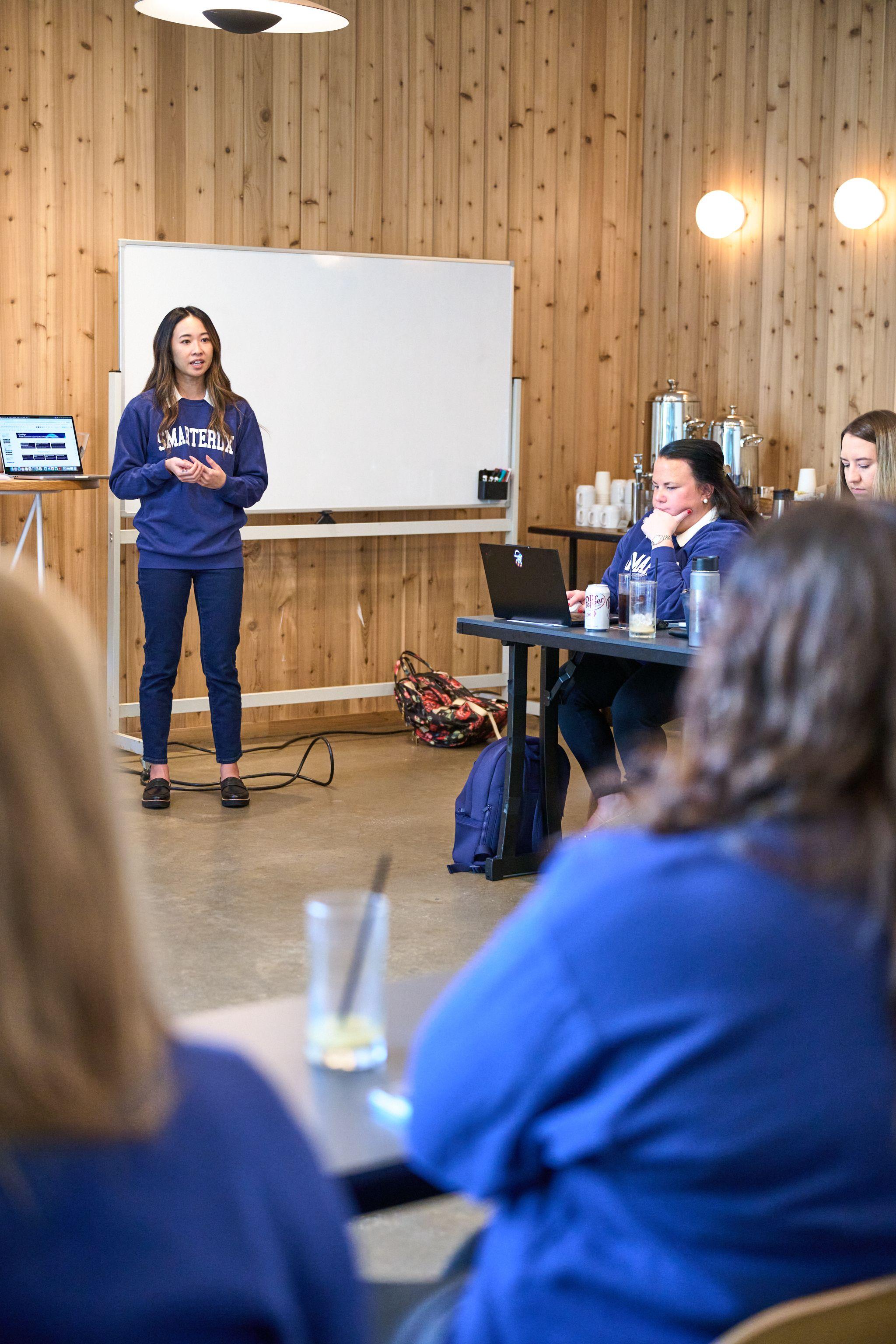 Woman in a company sweatshirt presenting at a whiteboard to a seated room — Team offsite photography, Austin A woman in a company sweatshirt presenting at a whiteboard to a seated room