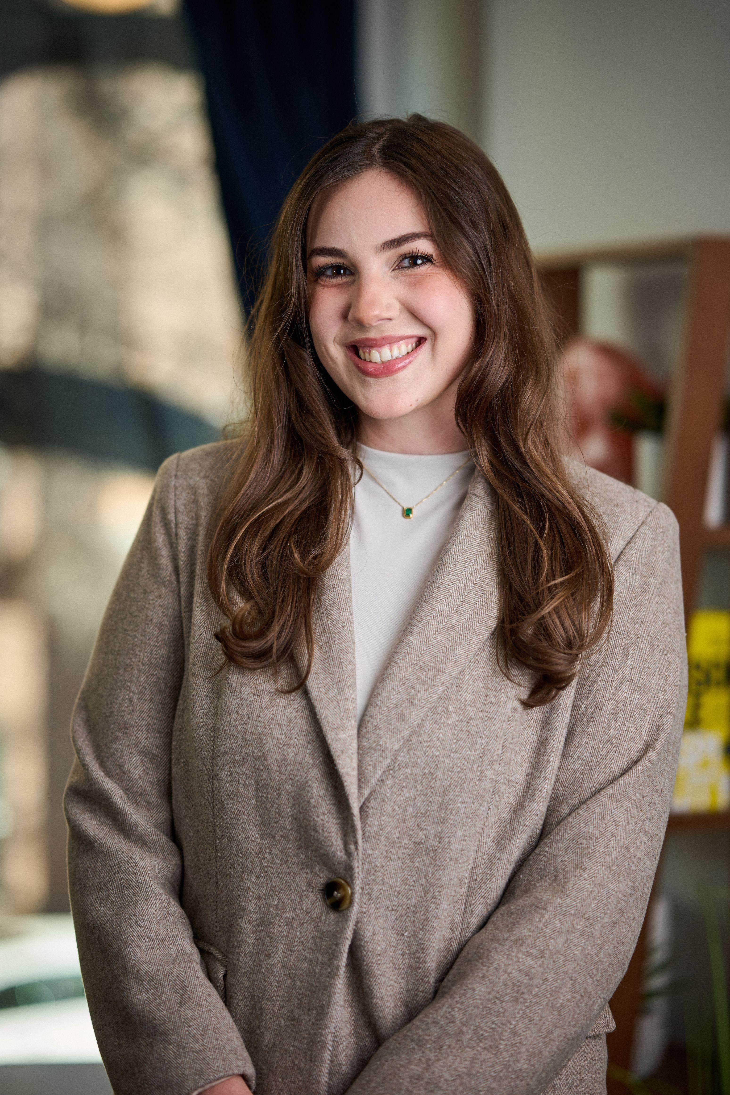 Young woman in a tan blazer smiling, a warmly lit interior blurred — Business headshots, Austin A young woman in a tan blazer smiling, a warmly lit interior blurred behind her
