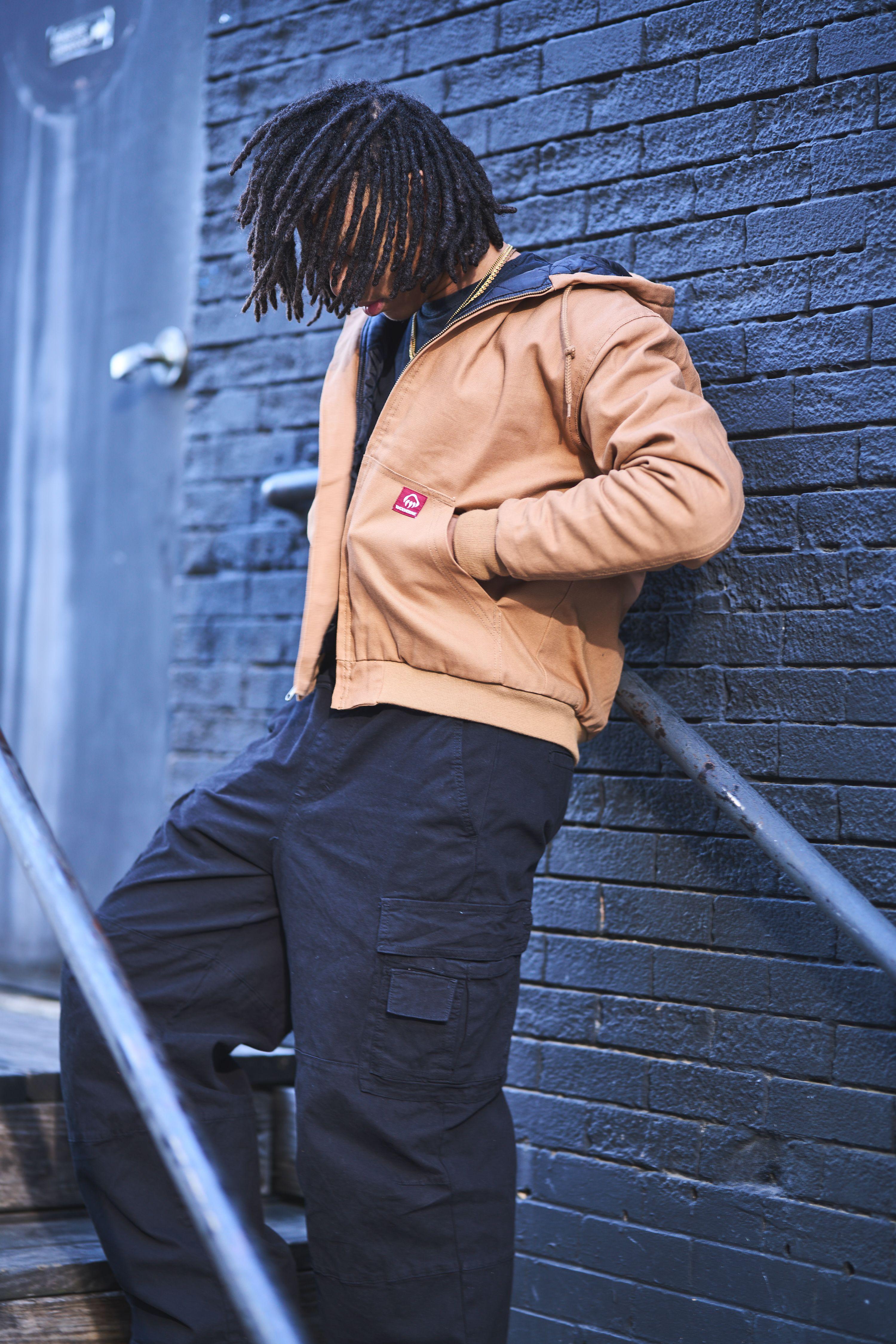 Model in a tan work jacket and dark trousers leaning against a painted brick wall — Fashion & editorial photography, Austin A model in a tan work jacket and dark trousers leaning against a painted brick wall.