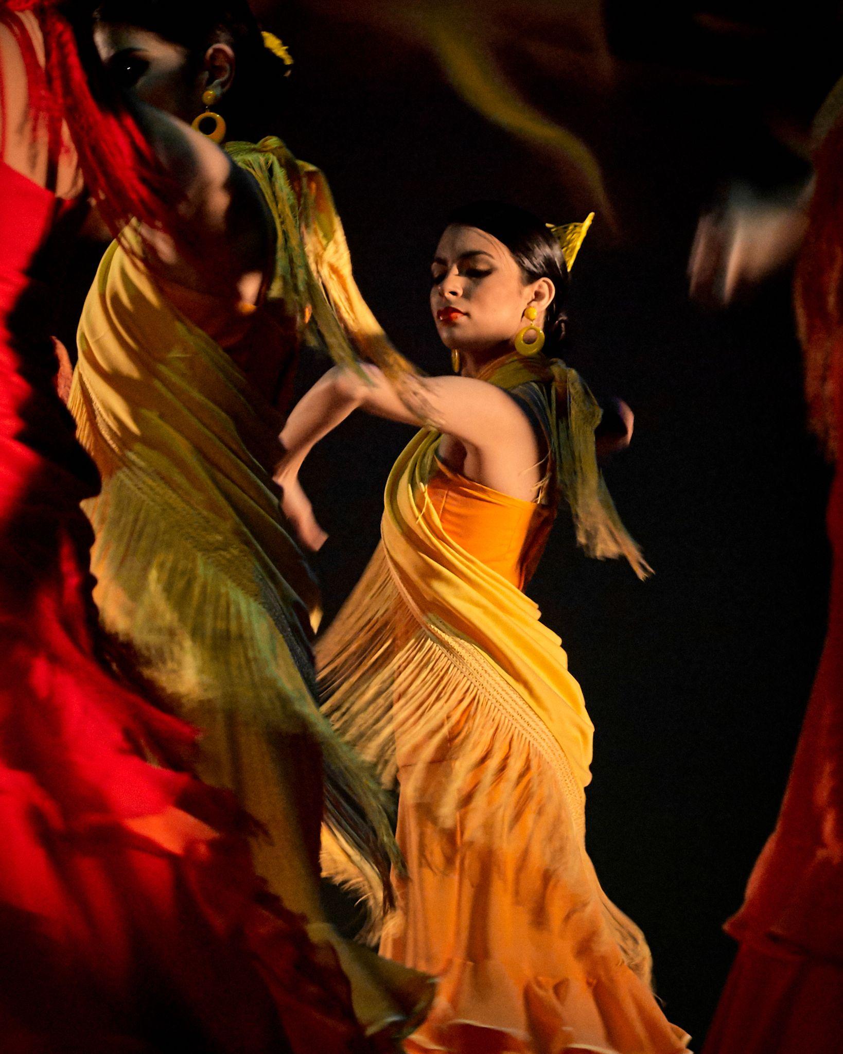 Two flamenco dancers mid-turn in yellow and orange dresses on a red-lit stage — Portrait photography, Austin Two flamenco dancers mid-turn in yellow and orange dresses on a red-lit stage.