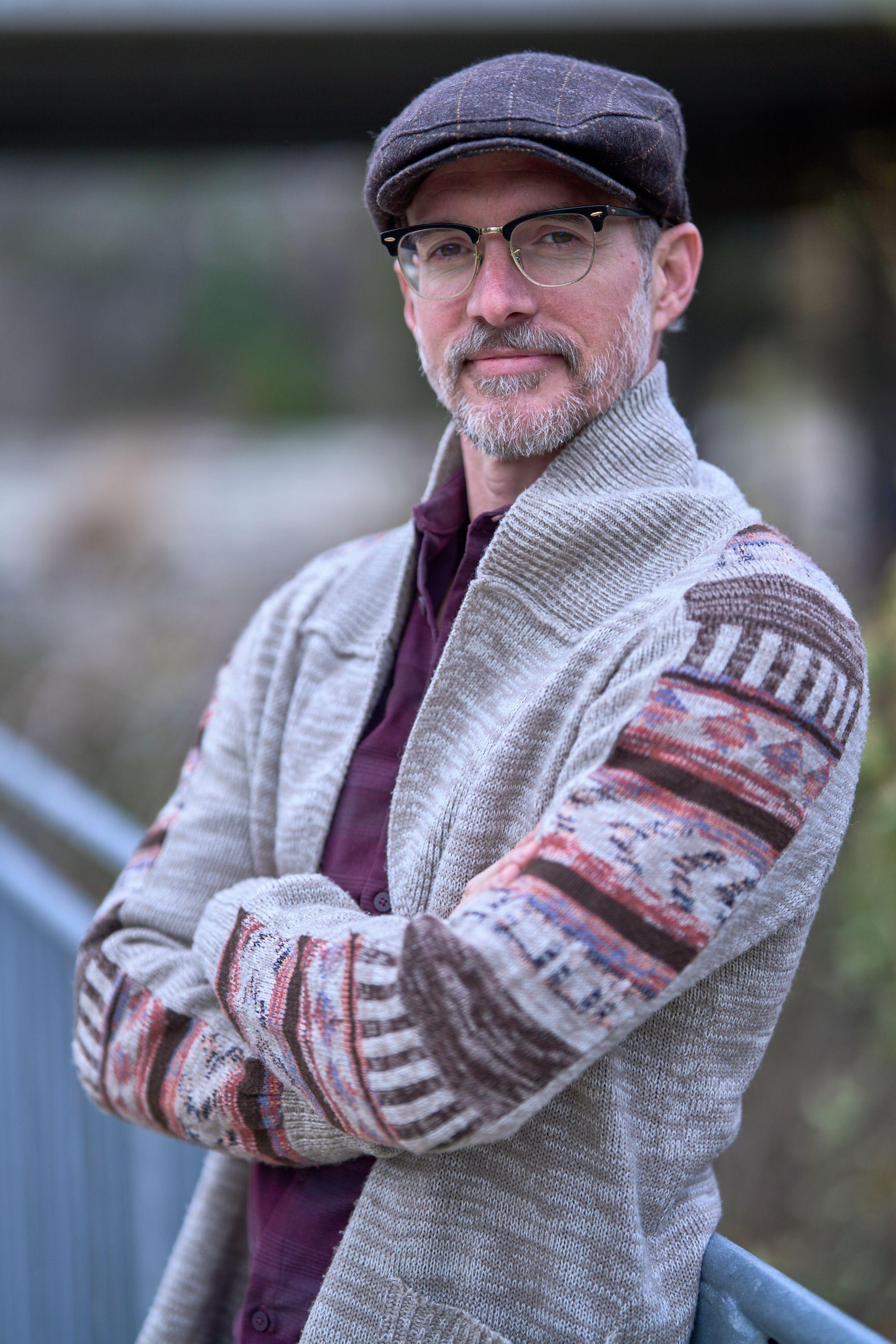 Man in a flat cap, glasses and patterned cardigan standing outdoors — Headshot photography, Austin A man in a flat cap, glasses and patterned cardigan standing outdoors.