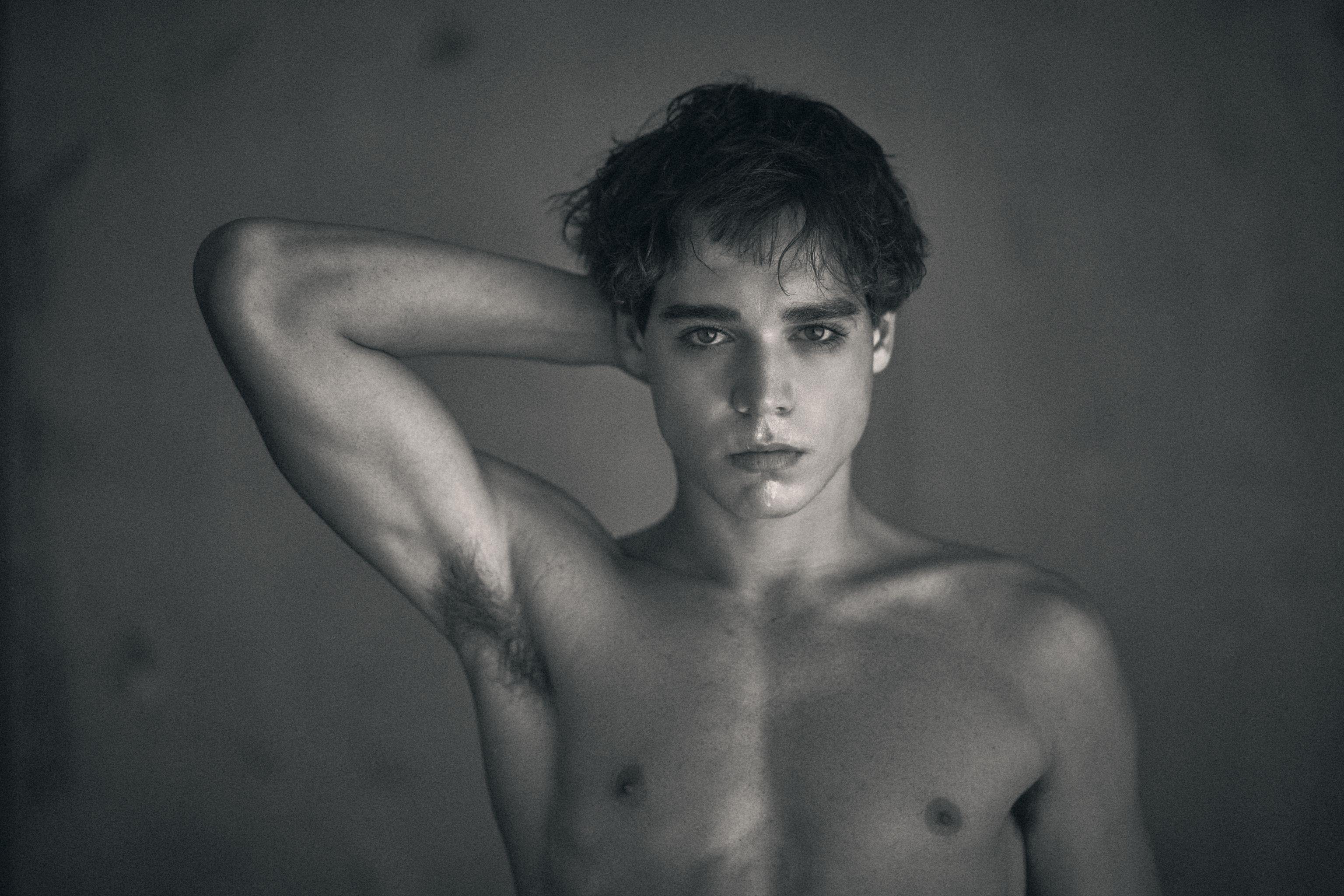 A black and white portrait of a shirtless young man with one arm raised behind his head