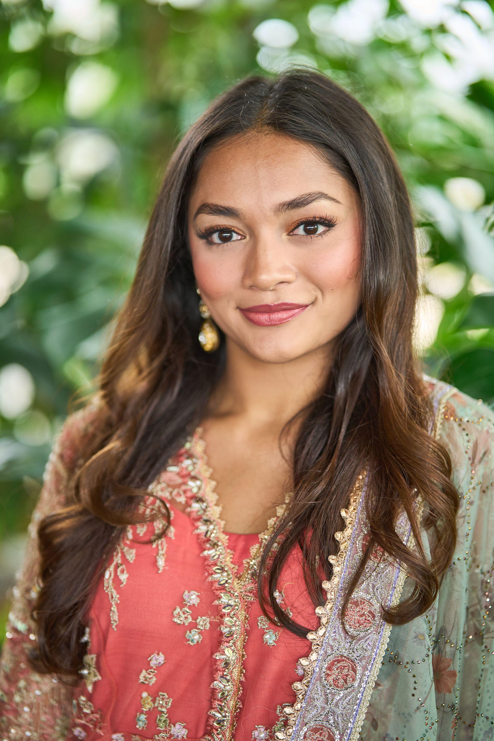 Woman in an embroidered coral outfit smiling among greenery — Headshot photography, Austin A woman in an embroidered coral outfit smiling among greenery.