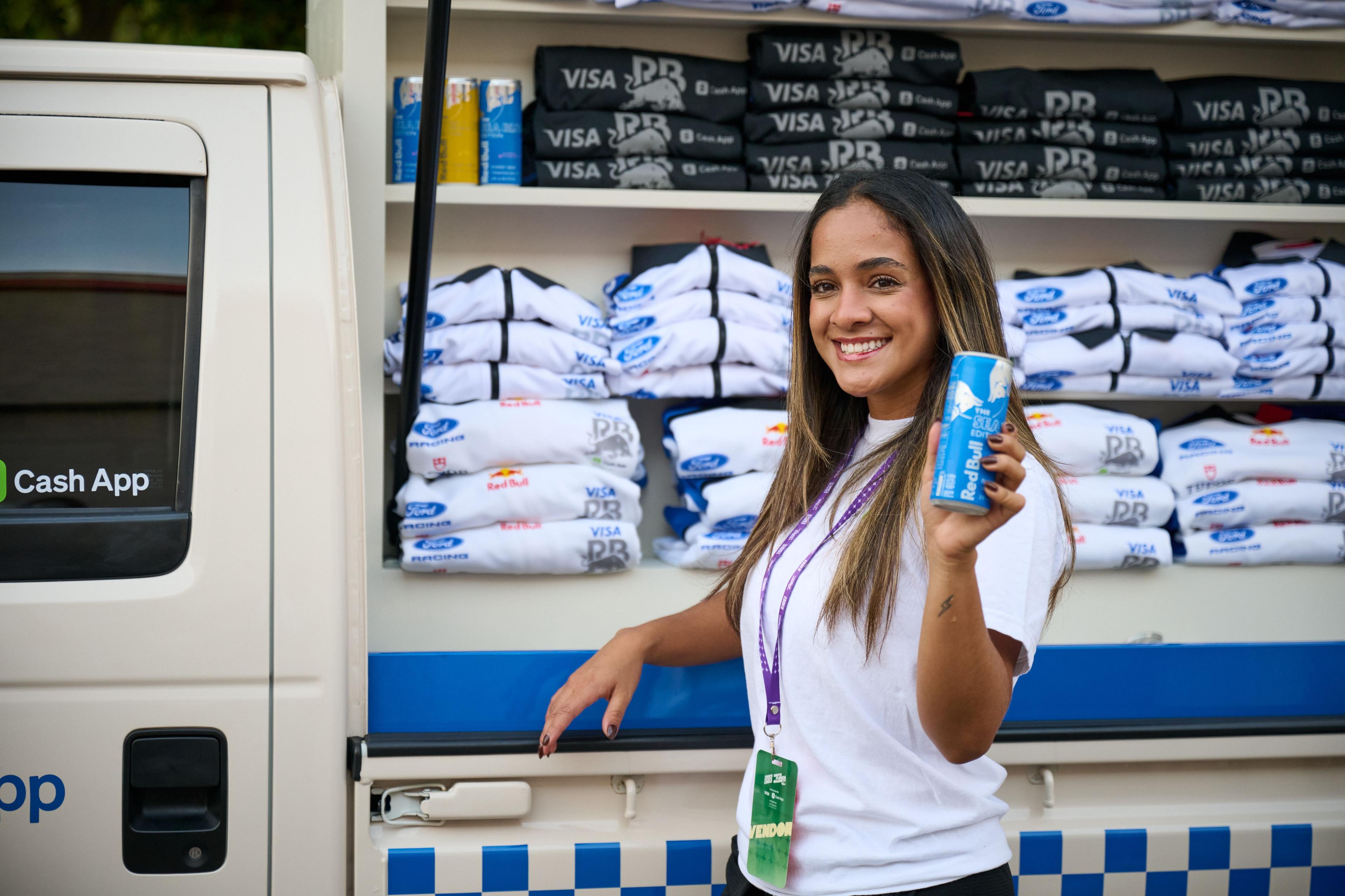 Brand host holding a can beside shelves stacked with Visa-branded tees and totes — Brand Collaborations photography, Austin A brand host holding a can beside shelves stacked with Visa-branded tees and totes.