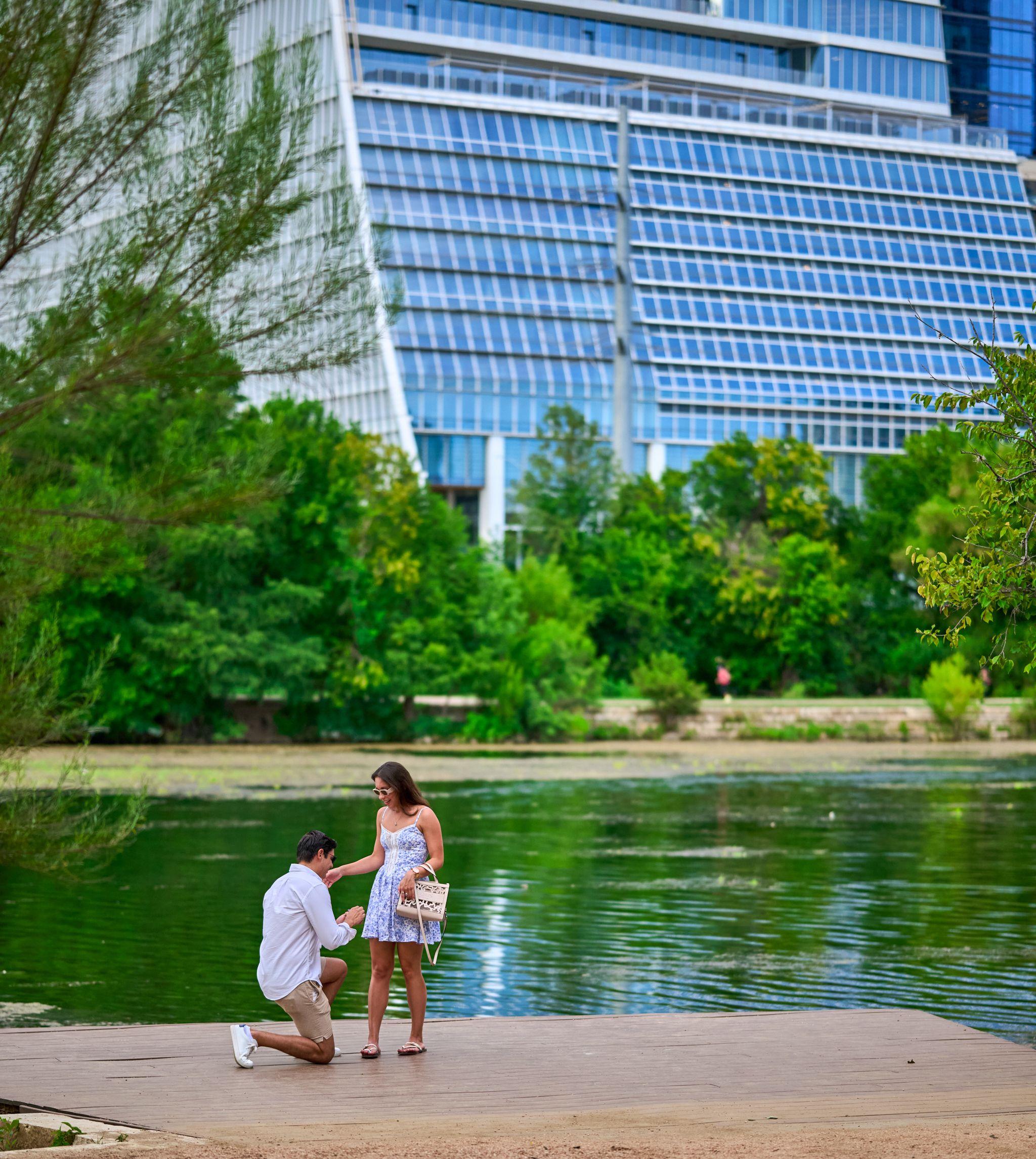 Distant view of a man kneeling to propose at the water's edge below a glass tower — Butler Metro Park photography, Austin A distant view of a man kneeling to propose at the water's edge below a glass tower.